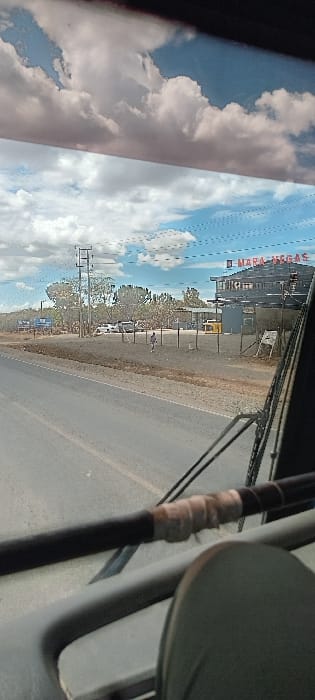 The image displays a roadside scene, likely captured from inside a vehicle looking out a window. The view is rotated 90 degrees clockwise from its natural orientation.

In the foreground, elements of the vehicle's interior are visible. On the left, a dark window frame borders the scene. On the right, a dark strap or pipe wrapped with light-colored tape and a curved, dark-colored object (possibly a mirror or part of a seat) are present.

The sky, dominating the upper portion of the view, is bright blue with scattered, fluffy white clouds, indicating daytime with fair weather. Reflections of these clouds can be seen on the window's surface.

Below the sky, a paved road extends horizontally across the frame, bordered by a dirt and gravel shoulder. A single person is seen walking on this dirt path towards the right side of the image. Several utility poles or support structures are spaced along the roadside. Beyond these, a line of trees and sparse vegetation is visible.

In the background, several buildings can be discerned. One prominent structure features a dark roof, and red lettering on its facade clearly spells out "MARA DEGAS." Other less distinct buildings and what appear to be parked vehicles are also present further along the road. The overall setting suggests a commercial or semi-urban area along a road, consistent with a location such as Narok Town ward in Kenya.
