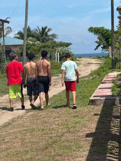 Four males are depicted walking away from the viewer along an unpaved dirt and grass path towards the ocean under a clear blue sky in Tela, Honduras. The individual on the far left wears a red t-shirt and neon yellow shorts with dark sneakers. The two central individuals are shirtless; one wears camouflage shorts and flip-flops, the other wears black shorts and appears barefoot. The individual on the far right wears a light blue t-shirt, red shorts, and dark sandals. To the left of the path, a building with a light blue corrugated roof is visible, along with a utility pole and dense tropical vegetation, including a palm tree. To the right, a raised concrete structure with reddish-brown top surfaces runs parallel to the path, partially shaded. The path leads directly to the visible blue expanse of the ocean, which is bordered by green foliage. The scene is illuminated by bright daylight, casting shadows on the right side of the path.