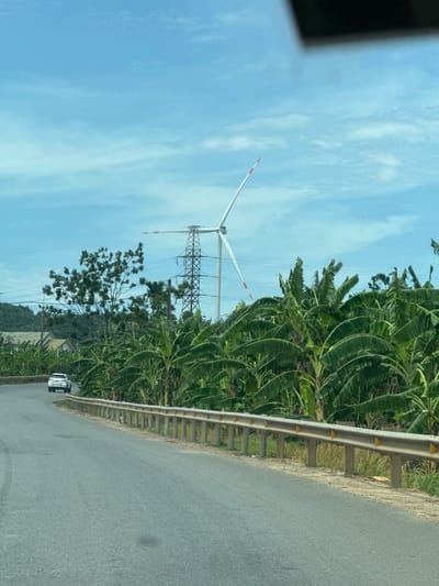This is a vertical, eye-level, outdoor, medium shot of a road in Tân Lập Commune, Vietnam. The road is paved and runs along the right side of the image. A metal guardrail with yellow reflectors lines the edge of the road. Lush green banana trees with large leaves dominate the left side of the scene, extending into the background. A white car is visible on the road, driving away from the viewer. In the distance, a wind turbine with white blades and red accents stands tall against a bright blue sky with scattered white clouds. A large electricity pylon with crisscrossing wires is also present near the wind turbine. The overall impression is of a rural landscape with a mix of natural and industrial elements under a clear sky, suggesting it is daytime. The scene appears to be peaceful and undeveloped.