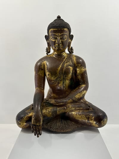 The image depicts a bronze statue of Buddha in a meditative pose, possibly situated in a museum or gallery, as it is placed against a white wall with an adjacent corner. The statue is dark bronze in color with visible traces of gold plating, giving it an aged appearance. Buddha is seated cross-legged with his hands resting in his lap, one over the other. He wears elaborate jewelry around his neck and dangling from his ears. His hair is styled in tight curls. The details of the face and body are sharply defined, suggesting a high level of craftsmanship. There is no indication of the statue's specific location in Lalitpur, Nepal, within the frame.