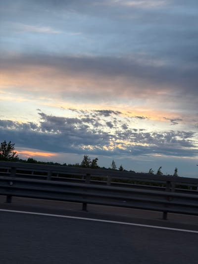 The image displays a scenic view of a road and sky, captured in Levashovo, Russia, likely during sunset or sunrise. The sky dominates the majority of the frame, showcasing a dramatic blend of clouds and colors. To the left, wispy clouds are illuminated by soft, warm golden and orange light, indicating the direction of the setting or rising sun. As the eye moves towards the right, the clouds become denser and darker, featuring various shades of blue, grey, and purple, with their edges still catching radiant golden highlights.

In the lower right portion of the image, a dark asphalt road is visible, featuring a clear white lane marking that extends towards the background. A metal guardrail runs horizontally across the mid-ground, acting as a divider between the road and the distant landscape. Below the guardrail, the dark silhouettes of trees and foliage are discernible, suggesting a natural environment bordering the road. The overall atmosphere of the scene is serene and tranquil, emphasizing the beautiful transitional colors of the sky during twilight. There is no visible text in the image.