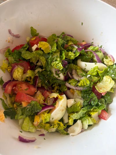 The image shows a salad in a white bowl. The salad consists of lettuce, tomato, red onion, and sliced hard-boiled eggs. The bowl appears to be indoors, possibly in a kitchen setting. The location is not evident from the image.