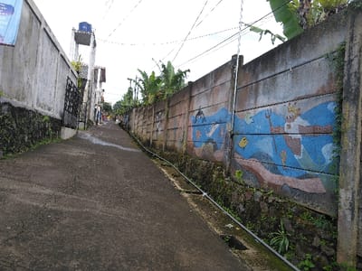 This image captures an upward-sloping, narrow concrete pathway in Hafjell, Norway, flanked by tall concrete walls under an overcast sky. The pathway, dark grey with visible cracks and wet patches, extends into the distance, curving slightly to the right.

On the left, a plain concrete retaining wall is visible, covered with moss and small plants at its base. Further up the incline on this side, a black metal gate is partially open, revealing a residential structure with an elevated water tank supported by concrete pillars. A light blue banner is partially visible at the far upper left.

On the right, a concrete panel wall extends along the pathway. This wall features a colorful mural depicting an underwater scene in blue and brown tones, showcasing a dolphin, a white bird, a yellow fish, and a merman-like figure holding a trident next to a castle. The base of this wall also exhibits moss and green vegetation. Above the right wall, large-leafed green plants are visible.

Overhead utility lines crisscross the sky. A thin, grey pipe runs along the base of the right wall, parallel to the pathway. No individuals or specific actions are discernible within the frame.