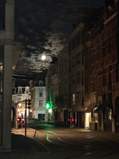 The image depicts a nighttime scene of a city street, identified as being in Ghent, Belgium, featuring a prominent full moon in a cloudy sky. The street is paved and has tram tracks that curve through the scene. Buildings line both sides of the street. On the left, a modern building with a light-colored facade is partially visible. Further down the street, older, multi-story buildings with traditional architecture are visible, some with illuminated windows. A traffic light with three circular lights is present on the left side of the street. On the right side of the street, a building displays a glowing green cross, indicating a pharmacy. Shopfronts with awnings and illuminated windows are also visible on the right. A tram or vehicle with red taillights is stopped on the left side of the street. The overall atmosphere is dark and atmospheric, illuminated by artificial lights and the moon.