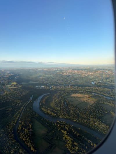 The image presents an expansive aerial view captured from an airplane window, looking down upon a diverse landscape under a clear sky. The horizon stretches across the middle of the frame, separating the deep blue sky above from the verdant ground below.
In the upper left portion of the sky, a faint, pale crescent moon is visible, accompanied by several thin, white contrails left by other aircraft. The sky transitions from a rich blue directly overhead to a lighter, slightly hazy blue and white towards the distant horizon, suggesting either early morning or late afternoon light. The right side of the view is partially obscured by the dark, curved frame of the airplane window.
The ground below is a tapestry of natural and developed features. Extensive dark green forests dominate much of the immediate foreground and mid-ground, densely packed with trees. A prominent, dark, winding river snakes through these forested areas, reflecting the sky in parts. Interspersed among the forests are agricultural fields, some showing distinct linear patterns, likely from cultivation, with long shadows cast across them by the low-lying sun. Beyond the immediate tree lines, the landscape opens up to reveal numerous scattered settlements, small towns, or villages characterized by clusters of buildings. These built-up areas extend towards the hazy horizon, where details become indistinct. Some larger structures, possibly industrial or commercial, are also visible.
The time of day appears to be either early morning or late afternoon, indicated by the warm, directional light and the long shadows stretching across the terrain. The weather is clear with some atmospheric haze in the distance. The context provided, Somma Lombardo, Italy, suggests this is a view over the Lombardy region, possibly showcasing parts of the Ticino River valley or similar features common in the Po Plain. No people or visible text are discernible within the frame.