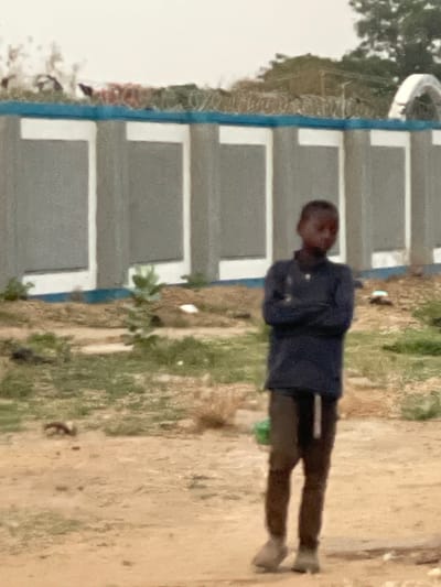 A person is standing outdoors in Hayin Banki, Nigeria, next to a building with a light-colored facade and blue trim. The person is wearing a dark blue shirt and dark pants. The ground is light brown and there is green vegetation present.