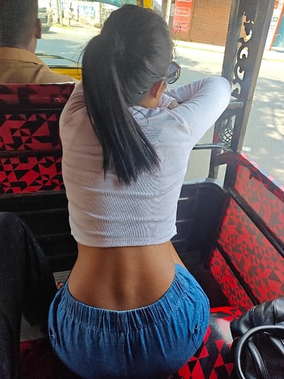 The image presents the interior of a motorized three-wheeled passenger vehicle (auto-rickshaw) on a sunlit urban street in Kochi, India. The primary subject is an individual viewed from the back, with long dark hair styled in a high ponytail. They are wearing a white, ribbed, long-sleeved crop top, which exposes the lower back, and blue elastic-waist pants. Sunglasses are positioned on the back of their head/neck. The individual is seated, leaning forward with their arms resting on the vehicle's structural frame. Ahead of them, the back of another individual wearing a light brown shirt is partially visible. The vehicle's interior upholstery, including the seats and side panels, features a repeating red and black geometric pattern. A black shoulder bag with a visible handle rests on the seat to the right of the primary subject. Through the vehicle's open side, a blurred background reveals a street, various buildings, and commercial signage, including a red sign, indicative of an urban setting.