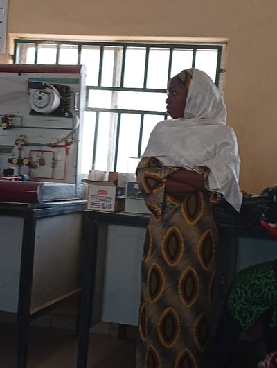 A woman in a patterned brown and gold dress with a white head covering stands in a room with industrial equipment. She has her arms crossed and is looking to the right. Behind her is a large window with multiple panes. To the left is a piece of industrial machinery mounted on a cabinet, and next to it are some boxes and equipment on a counter. Some fabric is visible on the right.