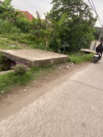 The image shows a paved road in Kecamatan Margaasih, Indonesia (latitude: -6.957, longitude: 107.548). A person riding a motorcycle is traveling away from the camera, to the right. The motorcycle is black with a red license plate. The person is wearing a black head covering. The road is bordered on the left by a dirt shoulder with a concrete drainage cover. The shoulder has green grass and shrubs, and there are some flowers visible. There are trees and bushes on the left side of the image in the background. The sky is overcast. There is a white building with a red roof in the far background.