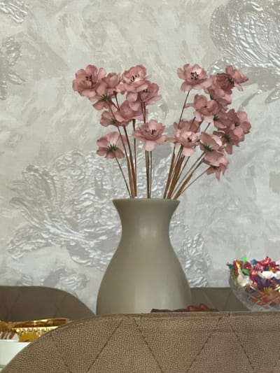 A bouquet of artificial pink flowers in a beige vase sits on a table in front of a textured wallpaper. To the right of the vase, a glass bowl filled with colorful candies is visible. In the foreground, a portion of a quilted brown surface is present. The image is a close-up shot, and no specific location in Sumgayit, Azerbaijan can be determined.