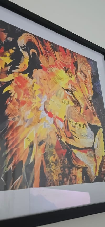 The image shows a framed collage artwork, predominantly composed of yellow, orange, red, and black tones. Fragments of text and images are discernible within the collage. Words like "intelligence," "fast," "forward," and "cuisine" are visible. The collage includes representations of faces and architectural elements. The artwork is framed in black with a white matte. The photograph appears to be taken indoors, possibly on a wall. There is no indication of Bergen, Norway or specific city context.