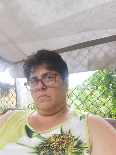 The image is a close-up, selfie-style portrait of a woman with short, dark hair and glasses. She is wearing a light green sleeveless top with a sunflower print. She is seated, with her head and shoulders filling most of the frame. Behind her, a green chain-link fence is visible, with foliage and trees beyond it. Above her, a light-colored canopy or awning provides shade. The lighting suggests it is daytime. The woman's expression is neutral, and she appears to be outdoors, possibly in a garden or backyard setting in Lomets, Bulgaria. There is no visible text in the image.