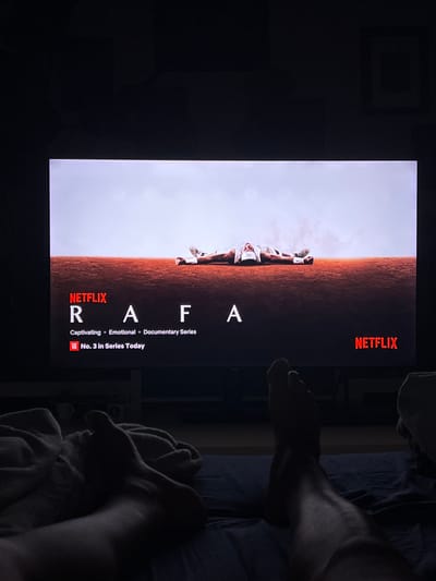 A Netflix screen displays a documentary series titled "Rafa," featuring a person lying on their back on a reddish-brown surface under a light sky. Below the title, descriptive text reads: "Captivating • Emotional • Documentary Series" and "TOP 10 No. 3 in Series Today." The Netflix logo appears in the bottom right corner. In the foreground, the legs and feet of a person are visible, lying on a dark blue surface, possibly a bed, covered by blankets. The scene is dimly lit, suggesting it is nighttime. There is no indication of the city of Mollet Del Vallès, Spain, in the image.