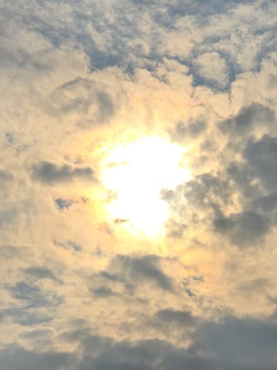 The image shows a cloudy sky with the sun shining brightly through the clouds. The clouds are white and gray, with some areas of blue sky visible. The sun is a bright, white orb in the center of the image, with rays of light emanating from it. The overall impression is one of a slightly overcast but bright day. The image is a photograph taken from below, looking up at the sky. There are no people, animals, or objects visible in the image. The location is described as Ермолаево, Russia, which is a city. However, the image itself does not contain any specific visual cues to confirm this location.