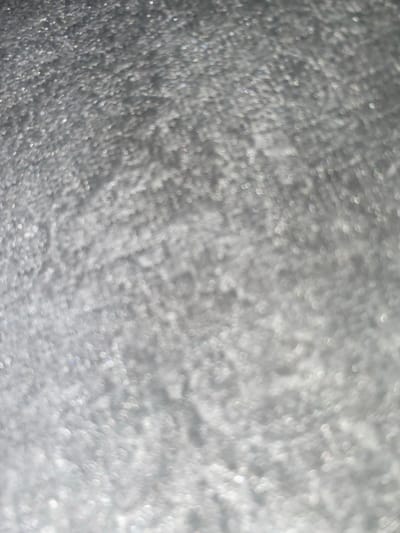 The image is an abstract, out-of-focus shot of a shimmering, granular surface, likely glitter or tiny reflective particles. The colors are predominantly shades of gray and white, with subtle hints of iridescence. There are no discernible objects, people, or environmental features that would allow for identification of a specific location, including Ablanitsa, Bulgaria. The texture appears to be densely packed and uniform across the frame.