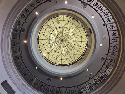 The image shows a downward view of a circular atrium with a geodesic dome ceiling. The dome is made of translucent panels divided by a dark metal framework, radiating from a central point. Surrounding the dome is a circular balcony with a decorative metal railing featuring ornate scrollwork. The balcony is illuminated by several circular lights embedded in the ceiling and wall. The walls of the atrium are painted in muted tones, and vents are visible on the walls. The overall impression is one of architectural elegance and symmetry.