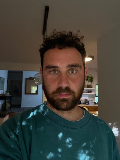 A man with curly brown hair and a thick, dark beard is wearing a teal sweatshirt with white bleach patterns. He is looking directly at the camera with a serious expression. The man is standing in a doorway, with a glimpse of a kitchen area and a living room beyond. The kitchen area contains white cabinets, wooden countertops, and a variety of kitchen supplies. The living room is visible in the background and includes a mirror and a rug. The scene is lit by natural light from an open window. The coordinates provided locate the scene in Alon HaGalil, Israel.