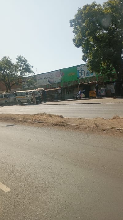 An urban scene presents a street with several vehicles parked along the curb. A building with a large sign reading "Mattresses & Cushions" is visible in the background, adjacent to a series of small market stalls. People are seen standing and sitting near the stalls. Trees are scattered along the top of the frame, partially obscuring the sky. The road occupies most of the foreground.