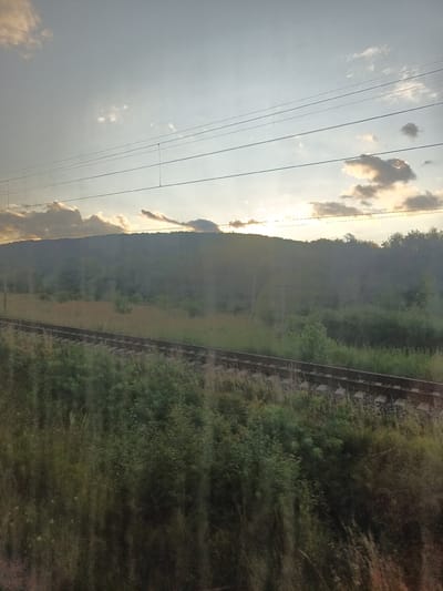 The image shows a rural landscape with a train track in the foreground. A tree-covered hill is visible in the background, with the sun setting behind it. The sky is partly cloudy, with the sun casting a warm glow. The overall scene is peaceful and serene.