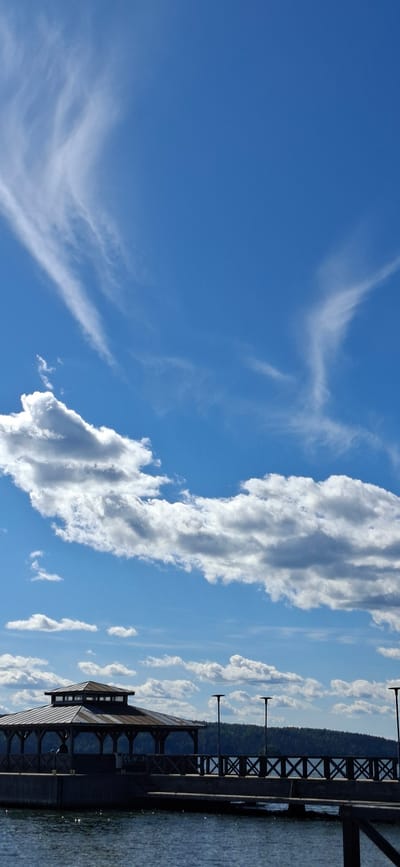 This image captures a serene scene under a bright blue sky filled with wispy white clouds and a prominent, elongated cloud formation. In the foreground, a modern, dark-colored structure with a multi-pitched roof and large windows is visible, situated on the edge of a body of water that reflects the sky. A wooden pier or walkway extends from the structure, featuring decorative railings with an "X" pattern and lampposts. The pier leads towards a densely forested hillside in the background. The water appears calm with subtle ripples, suggesting a peaceful atmosphere. The lighting and the clear blue sky indicate it is daytime, likely late morning or early afternoon. The presence of the pier and the building suggest a recreational area or a waterfront establishment. The scene evokes a sense of tranquility and natural beauty, typical of a sunny day in a lakeside or seaside location. The image appears to be a static shot with no discernible activity taking place.