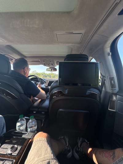 The interior of a vehicle, possibly a luxury car, is shown from the rear passenger perspective. The driver, a man with short dark hair and glasses, is seen looking at the road ahead and driving the vehicle. Two water bottles are visible on the center console. A display screen is mounted on the back of the driver's seat. The upholstery is light gray. Part of a person's legs and feet are visible in the right side of the photo. The setting is most likely the city of Ravenna, Italy, based on the request.