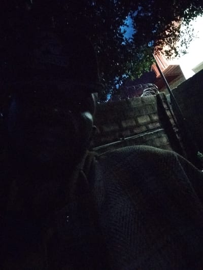 The image depicts a dark, low-light scene, likely taken at night or in a dimly lit indoor space. The primary subject appears to be a person, or at least part of a person's head and shoulder, visible on the left side of the frame. They are largely obscured by shadow, making specific details difficult to discern. A textured fabric, possibly a blanket or clothing, covers a significant portion of the lower right side of the image, suggesting the person is draped in it or resting.
In the background, there are hints of an outdoor setting. A brick wall is visible, with what appears to be barbed wire or a similar protective fencing material stretched across its top. Above the wall, foliage from trees is silhouetted against a bright, possibly artificial light source, which also illuminates a portion of a building with a reddish-brown facade. The sky above the trees shows a deep blue, suggesting it is nighttime.
The overall mood is somewhat mysterious and intimate, due to the darkness and the close-up perspective. There is no visible text in the image. The location context of Mangu Town, Nigeria, is noted but not visually evident from the image itself. The activity is unclear, but it seems to be a moment of rest or quiet observation.