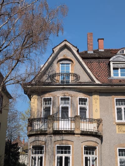 The image displays the upper sections of a multi-story residential building located in Heidelberg, Germany. The structure features a grey stucco facade with decorative architectural elements, including molded window surrounds and a yellow trim accenting a prominent arched window within a triangular dormer. The roof is pitched and covered with reddish-brown tiles, from which two red brick chimneys protrude.

A small, curved wrought-iron Juliette balcony is situated directly beneath the arched dormer window. Below this, a larger, curved balcony with a black wrought-iron railing and stone supports extends from a corner of the building. Numerous windows with white frames are visible across the facade; some are multi-paned.

The environment includes a clear blue sky overhead. A large, deciduous tree with bare branches extends into the upper left quadrant of the frame. Portions of an adjacent building with a lighter-colored facade are visible to the left, and distant rooftops and vegetation can be observed in the lower-left background. Sunlight illuminates the scene, casting shadows consistent with a light source from the right. There are no individuals, specific actions, or interactions depicted.
