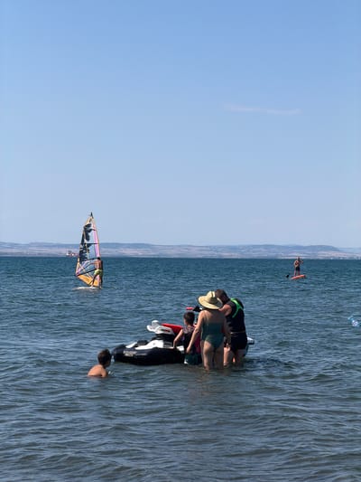 In Atiya, Bulgaria, a group of people are enjoying a sunny day at sea. In the foreground, a family stands in the shallow water next to a personal watercraft. A woman in a green swimsuit and a straw hat stands facing away from the camera, while a man in a life vest and shorts stands beside her, also facing away. A young boy in a dark swimsuit stands near them. In the middle ground, a windsurfer with a colorful sail navigates the waves, and in the distance, a person stands on a paddleboard. The sea is calm with gentle ripples, and the sky is a clear, bright blue. A cargo ship is visible on the horizon, near the coastline.