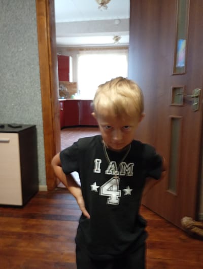 A young boy with blond hair stands in a room with wooden floors. He is wearing a black t-shirt with white lettering that says "I AM 4" and two white stars. The boy has his hands on his hips and is looking at the camera with a serious expression. Behind him, a doorway leads to another room with red cabinets and a window. A wooden door is on the right side of the image. The scene appears to be indoors, possibly in a home.