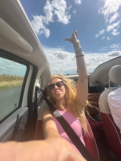 A woman with long blonde hair and sunglasses, wearing a pink top, is in the passenger seat of a convertible car. Her left arm is raised in the air, and her hand is in a gesture. She is puckering her lips. The car is moving, as indicated by the blurred scenery outside the window, which shows a road and dry vegetation. The sky is blue with scattered clouds. The image appears to be taken in Alaincourt, France, based on the surrounding landscape which is consistent with the region.