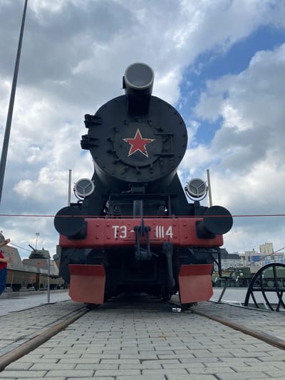This is a frontal, eye-level, medium shot of a black steam locomotive with a red front bumper, located in the Museum of Military Equipment in Verkhnyaya Pyshma, Russia. The locomotive has a large red star with a white outline on its front. Two circular headlights are mounted above the bumper. The railway tracks in front of the locomotive are made of large grey paving stones. Behind the locomotive, several tanks and military vehicles are parked, some partially obscured by the locomotive itself. In the background, there are buildings and a cloudy sky with patches of blue. A red rope barrier is visible in front of the locomotive, and to the far left, a person in a red shirt and blue jeans is partially visible, looking at the tanks. The overall environment appears to be an outdoor museum or exhibition space.
