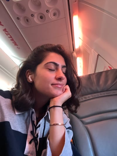 The image shows a young woman with her eyes closed, resting her head on her hand. She is wearing earbuds and a bracelet. The background suggests she is in an airplane cabin, with overhead lights and seats visible. The text "MALTA" is visible on the wall of the cabin, indicating the airline or destination. The lighting is dim and reddish, consistent with cabin lighting. The woman appears relaxed and possibly asleep or dozing. The location in the city of Lonate Pozzolo, Italy, is likely the Malpensa Airport, which serves the Milan metropolitan area and is the principal airport for Malta Air flights.