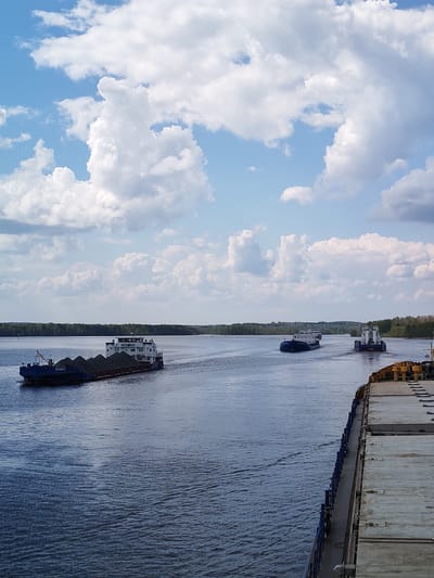 The image shows a wide river, likely the Oka River, with several barges navigating it. The perspective is from another barge, possibly anchored or moving slowly. In the foreground, the deck of this barge is visible, with a section of its structure and cargo or equipment in yellow tones.
Three other barges are visible on the river. The barge closest to the left edge of the frame is loaded with a dark material, possibly coal, and is being pushed by a tugboat. Two more barges are further down the river, moving away from the viewer and also being pushed by tugboats.
The water is slightly choppy with ripples and wakes indicating the movement of the vessels.
The horizon is marked by a line of trees, suggesting a rural or semi-rural location.
Above, the sky is predominantly blue with scattered, puffy white clouds.
Based on the location, the barges are likely engaged in the transportation of goods or materials along the river.