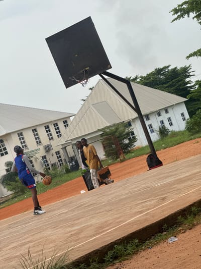 The image depicts an outdoor basketball court with two buildings in the background, situated in Amansee, Nigeria. In the foreground, a young person wearing a blue basketball jersey and shorts is dribbling a basketball. They are also wearing a headscarf and white sneakers. To the right of the dribbler, two other individuals are standing. One is wearing a yellow shirt and patterned pants, holding a briefcase. The other person, wearing a white shirt and black pants, is partially obscured. A basketball hoop with a black backboard and a red net is visible, tilted at an angle. The court is made of concrete with visible lines, and the surrounding area is covered in reddish-brown dirt and patches of green grass and trees. The sky is overcast. The buildings in the background are modern with white exteriors and multiple windows. One building has a sign that reads "NEEDS ASSESSMENT FUND PROJECT". A black backpack with red accents is placed on the dirt near the base of the basketball hoop's pole.