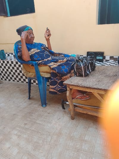 An elderly individual is situated outdoors in Ufuma, Nigeria. The subject, likely female, is seated in a faded blue plastic chair cushioned with a light brown pad. She wears a dark blue headwrap with a teal band and a dark blue dress patterned with gold and lighter blue motifs. She holds a dark-colored smartphone in her right hand, looking at its screen, while her left hand is raised near her temple. Her legs are propped onto a small, rectangular, worn wooden table. A black plastic bag is positioned on the table surface. Beneath the table, a portion of a black and silver walking aid is visible on the concrete ground, near her right foot, which is shod in a white patterned flip-flop. The environment features a light gray concrete floor and a plain light beige wall in the background. A section of the lower wall to the left exhibits black and white checkerboard tiles. A dark electronic device, resembling a radio or speaker, rests on the ground against the beige wall behind the table. A dark teal plastic jug is visible on the tiled wall section. Natural daylight illuminates the scene, with a pronounced orange lens flare in the lower right foreground.