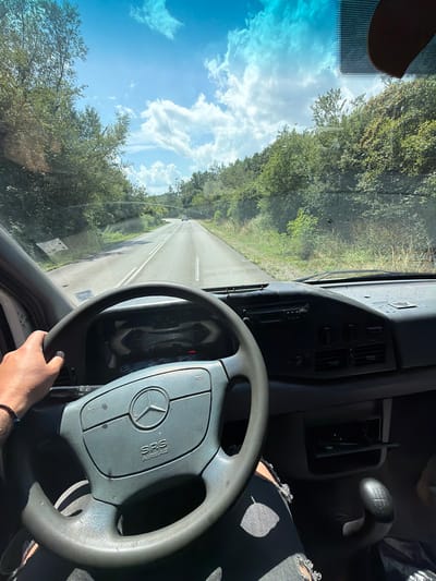 The image shows the interior of a Mercedes-Benz van, with the driver's hand on the steering wheel as the vehicle is in motion. The steering wheel displays the Mercedes-Benz logo and "SRS AIRBAG". The dashboard features a radio and climate controls. The road ahead is paved with white lane markings and is bordered by lush green trees and vegetation. The sky is a bright blue with scattered white clouds. There are no visible elements that would specifically indicate the location as Garnia, Bulgaria. The scene depicts a typical driving experience on a sunny day.