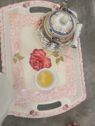 A high-angle, close-up shot captures a decorative tea tray, likely made of plastic, resting on a grey, concrete surface. The tray itself features a pink floral pattern against a cream background. Situated centrally on the tray is a white teacup filled with a golden-yellow liquid, presumably tea. To the upper right of the cup is a large, red floral design. Dominating the upper portion of the tray is a vibrantly decorated teapot. The teapot boasts intricate patterns of various colors, including blue, red, yellow, and green, with a light-colored knob on its lid. A metallic handle and spout are visible. The scene appears to be set outdoors, given the concrete ground and the natural lighting. The presence of a bare foot, with visible toes, at the bottom right of the frame suggests a person is present, though only partially visible. The overall impression is one of a traditional tea setting in what could be an outdoor urban or semi-urban environment. The location is identified as Kan Mehtarzai, Pakistan.