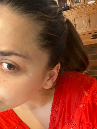 The image displays a close-up, angled view of an individual's right side, identified as being in Ancenis, France. The subject's right eye, with a visible brown iris and dark pupil, is in the lower left quadrant. The forehead, part of the nose bridge, cheek, and right ear are visible. The skin tone is fair with minor imperfections on the forehead and cheek. Dark brown hair, appearing to be pulled back into a ponytail, frames the face and ear; lighter brown strands are discernible within the ponytail. The subject is wearing a bright red garment, likely a top or dress, featuring patterned eyelet cutwork on the visible shoulder and upper arm. The background is out of focus, revealing a light-colored wooden cabinet with multiple drawers and wooden knobs in the upper right. Below the cabinet, a dark horizontal surface, possibly a table or counter, is visible, alongside a reddish-brown textured surface resembling brickwork.