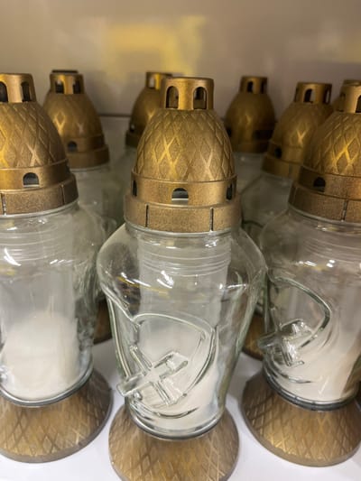 This media file shows a close-up, vertical view of a row of decorative glass candle holders. The objects are arranged in a staggered fashion, with the tops of some overlapping the bottoms of others. Each candle holder consists of a clear glass container with a metallic gold-colored cap and base. The caps are intricately designed with a diamond-shaped pattern and have openings for airflow. The bases are also circular and share the same textured gold finish.

Inside the glass containers, white candles are visible. Some of the glass jars have a raised cross shape molded into the glass, suggesting a religious or commemorative purpose. The lighting appears to be artificial, with a soft glow reflecting off the metallic surfaces and glass. The background is a plain, light-colored surface, possibly a shelf or wall, which helps to isolate the objects. The scene is static, with no discernible activity other than the presentation of the objects themselves.

There are no people visible in the media file. The setting is likely a retail environment or a display of home decor items. Given the location context of Ivanivka, Ukraine, these candle holders might be associated with local customs or memorial traditions. No visible text is present in the image. The overall impression is one of solemnity and remembrance, evoked by the design and material of the candle holders.