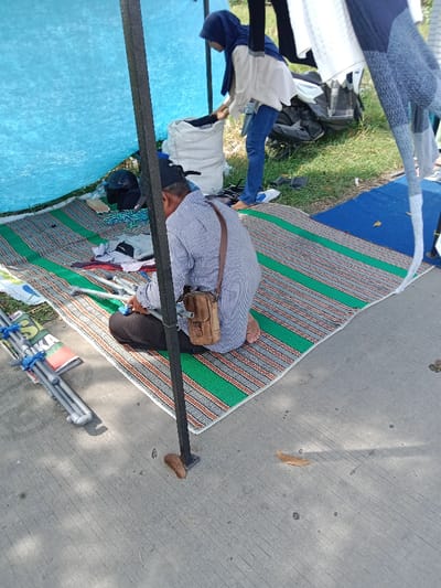 The image depicts a scene from a street market in Kendal, Indonesia, specifically at the coordinates -6.935 latitude and 110.203 longitude. A man, wearing a blue and white patterned shirt and a brown leather bag, sits on a green and orange striped mat. He is holding a gray metal pipe, seemingly part of a tent structure. A woman, wearing a white headscarf and jeans, is standing nearby, out of focus, and appears to be organizing items under a blue tarp. Another blue tarp is partially visible in the background. The scene is situated on a paved concrete surface with green grass and a blue mat nearby. The man's posture suggests he might be waiting or resting while the woman is engaged in setting up the market stall. The scene captures a moment of everyday life at a bustling Indonesian market.