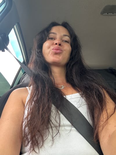 The image shows a woman in the driver's seat of a car, with her seatbelt fastened. She is looking at the camera and puckering her lips. The car appears to be parked, possibly in Slyudyanka, Russia, given the context. The woman has long, dark hair and is wearing a white tank top. The interior of the car is dimly lit, with natural light coming in from the side window. There is a black bag visible in the background. The overall impression is of a casual selfie taken while in the car.