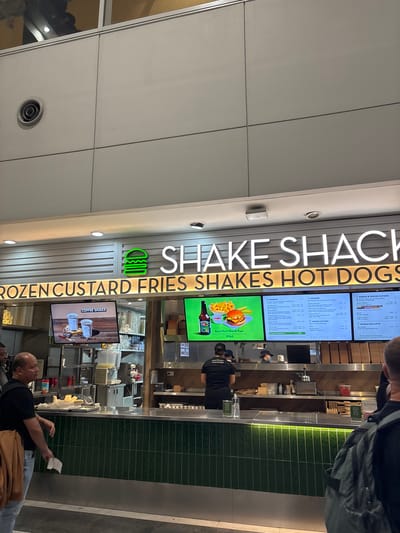 The image shows the interior of a Shake Shack restaurant, likely located in Pasay, Philippines, given the price points visible on the menu. The prominent signage reads "SHAKE SHACK" in large white letters, with the iconic green burger logo to the left. Below this, a backlit sign displays "ROZEN CUSTARD FRIES SHAKES HOT DOGS".
Several menu boards are visible. One to the left displays "COFFEE SHAKE" with images of two coffee shakes. Another central menu board shows categories like "BURGERS," "CHICKEN," and "CRINKLE CUT FRIES," with prices in what appears to be Philippine Pesos (P). To the far right, another menu lists "SHAKES & FROZEN CUSTARD" and "MANILA EXCLUSIVE" items.
In the foreground, a man wearing a black t-shirt and light-colored jeans stands to the left, holding a white slip of paper. He has a brown jacket draped over his left shoulder. Further to the right, another person with a backpack is partially visible, facing away from the camera.
Behind the counter, staff members are visible working. One person in a black uniform with "SHAKE SHACK" on the back is preparing orders. Another person, wearing a mask and a black cap, is also behind the counter. The counter area features stainless steel surfaces, green tiled walls below, and shelves stocked with various items. A large dispenser of yellow liquid is also present on the counter.
The environment appears to be a busy indoor establishment, possibly an airport or a mall, given the modern architectural elements of the ceiling and walls. The lighting is a mix of overhead spotlights and the backlit signage.