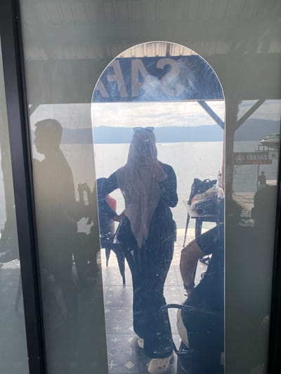 A vertical, full-body mirror reflects a young woman wearing a hijab and sunglasses. She is standing and taking a selfie. The woman is wearing a loose black outfit and light-colored clogs. In the reflection, a table with bags and some objects sits behind her. The background of the reflection shows a body of water with hills in the distance, suggesting a lakeside or coastal location. Through the mirror and the window, a shadowed silhouette of another person can be seen to the left, facing away from the mirror. To the right of the reflection, a sign with the word "SAFARI" is visible, indicating a business or attraction. The floor beneath the woman in the reflection is tiled with a diamond and cross pattern. The overall scene suggests an outdoor setting, possibly a cafe or a scenic spot in Tuk Tuk, Samosir Island, Indonesia, known for its lakeside views and tourist activities.