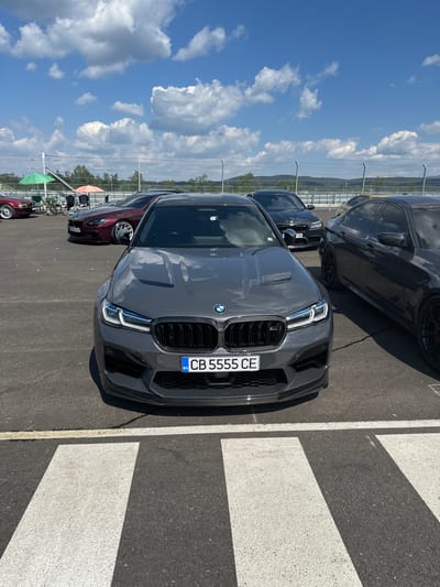 The image captures a frontal view of a dark gray BMW M5 on a paved surface, likely a racetrack or parking lot, at the city of Dospey, Bulgaria. The car is positioned in the foreground, with its license plate reading "CB 5555 CE" clearly visible. Behind it, several other vehicles are parked, including a burgundy BMW and two dark sedans. In the background, a wire fence separates the parking area from a vegetated landscape with rolling hills under a bright blue sky dotted with white clouds. Two green and orange umbrellas are set up near some chairs, suggesting a break area. The ground is asphalt with white markings, indicating a designated area or lane.