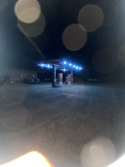 The image depicts a gas station at night in Sasaro, Nigeria. A canopy is centrally positioned, illuminated by a linear array of eight visible cool-toned lights. The partial signage on the canopy reads '...CO PETROLEUM'. Two gas pumps, colored orange-red and gray, are beneath the canopy on the right. To the left, a dark building, presumably a station office, displays a single illuminated window. Several indistinct objects, possibly covered vehicles or equipment, are discernible in front of the building. The ground is unpaved. The scene is devoid of people. The image is significantly affected by multiple large, blurred circular light artifacts, and a bright orange-yellow light flare originates from the lower-left foreground.
