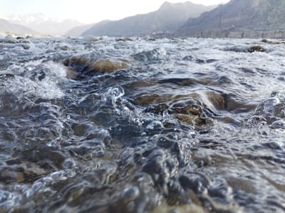 The image displays a low-angle, close-up perspective of a flowing river or stream, located in Qoraqiya, Uzbekistan. The foreground is dominated by clear, turbulent water characterized by ripples, small waves, and splashes. Submerged rocks and pebbles of brown and grey tones are visible through the water's surface. Several distinct water droplets are momentarily suspended above the surface, indicative of active flow. The midground shows the continuous water body extending into the distance. The background features a rugged, mountainous landscape under a bright, clear sky. Multiple mountain ranges are present, with distant peaks appearing lighter, consistent with snow cover or strong illumination. No individuals, man-made structures, or artificial objects are visible within the frame. The scene is illuminated by natural daylight.
