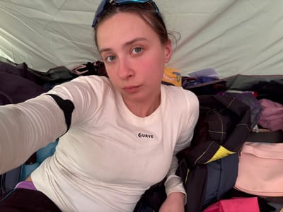 A young adult female with blue sunglasses on her forehead, wearing a long-sleeved white shirt with the word "Curve" on it, lies inside a tent. The tent is filled with various items of clothing, including pink, black, and yellow jackets, and a pink bag. The image was taken at an angle, looking down at the subject.
The location is not specified in the image.