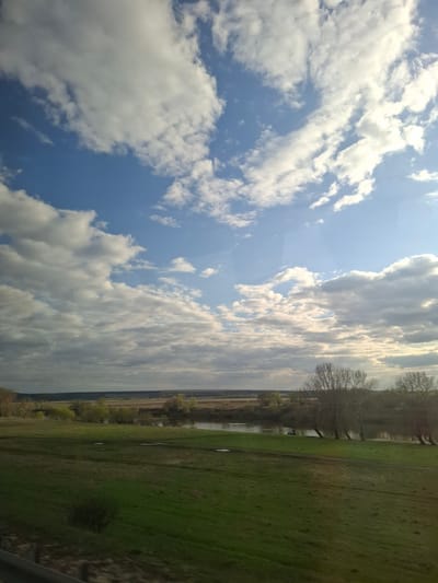 The image captures a serene, expansive landscape under a dynamic sky, characteristic of a rural or semi-rural setting in Knyazevo, Russia. The sky dominates the upper two-thirds of the frame, presenting a clear blue expanse dotted with numerous white and light grey cumulus and stratocumulus clouds. Bright sunlight is visible, illuminating the clouds from behind and casting a glow through them.

Below the horizon, the land unfolds with a broad field of green grass in the foreground, indicating lush vegetation. A narrow body of water, likely a river or stream, runs horizontally across the midground, its surface partially reflecting the sky. Along the far bank of this water feature, a line of bare, deciduous trees stands, their branches devoid of leaves, suggesting either late winter or early spring. Further in the distance, the landscape recedes to a faint, slightly hazy horizon line, with more distant trees and a seemingly more overcast sky.

The time of day appears to be mid-morning or afternoon, given the bright, direct sunlight. The weather is partly cloudy with significant sun. There are no people or specific activities depicted. The overall impression is one of peaceful natural beauty and vastness. No text is visible in the image.