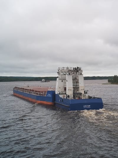 A blue and orange cargo ship, identified as "АЛЬТАИР" (Altair) from Saint Petersburg, Russia, travels on a wide body of water under a cloudy sky. The vessel consists of a flat barge section and a multi-level control tower structure. The water surface is moderately rough with visible wake from the ship's movement. In the background, wooded shorelines can be seen on both sides of the water, with another distant vessel visible in the center. The photograph does not confirm it being located in Анхимово, Russia, but this information is given in the prompt.