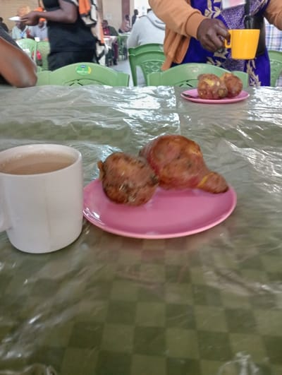 This image depicts an indoor dining or communal eating area, likely in Riara Ridge, Kenya. In the immediate foreground, a white mug filled with a light brown beverage, possibly tea or coffee with milk, is placed on a table. Beside it, a pink plate holds two large, irregularly shaped, reddish-brown cooked root vegetables, resembling sweet potatoes or yams. The table is covered with a clear plastic sheet over a green and white checkered tablecloth.
In the mid-ground on the right, a person, whose lower arm and torso are visible, wears a blue patterned garment and an orange long-sleeved top. They are holding a bright yellow mug. Below their hand is another pink plate, also containing two similar cooked root vegetables.
The background shows several people, mostly out of focus. Some are seated at tables with bright green plastic chairs, while others appear to be standing. The setting suggests a functional hall or large dining area, possibly for a school, community center, or similar establishment. The activity taking place is people gathered for a meal or refreshments, with the prominent food items indicating a simple, possibly breakfast or snack, offering. No specific time of day is discernible, and no visible text is legible. The atmosphere appears calm and routine.